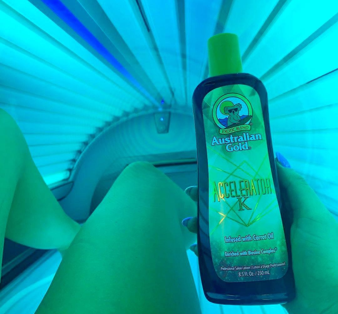 australian gold tanning lotion accelerator k is being held in a laydown sunbed. you can see the tanner's legs and their manicured right hand that is holding the tanning lotion in the sunbed